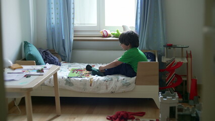 Candid little boy trying to put socks sitting in bed inside bedroom. Upset emotion of child...