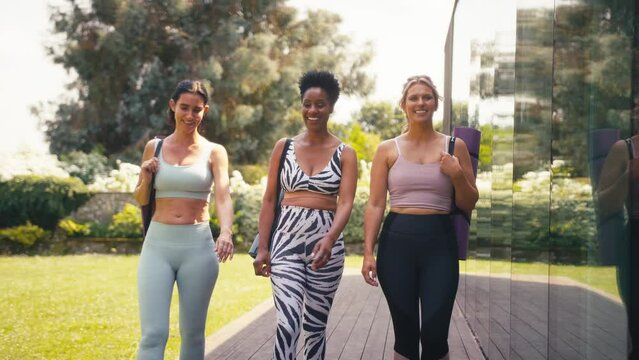 Three mature female friends wearing fitness clothing carrying exercise mats outside gym or yoga studio - shot in slow motion