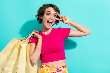 Portrait of cheerful pretty lady hold store bags demonstrate v-sign near eye isolated on turquoise...