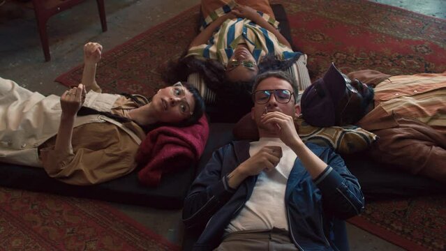 High angle shot of young Black and Caucasian squatters lying on mats on floor in abandoned house smoking weed and talking