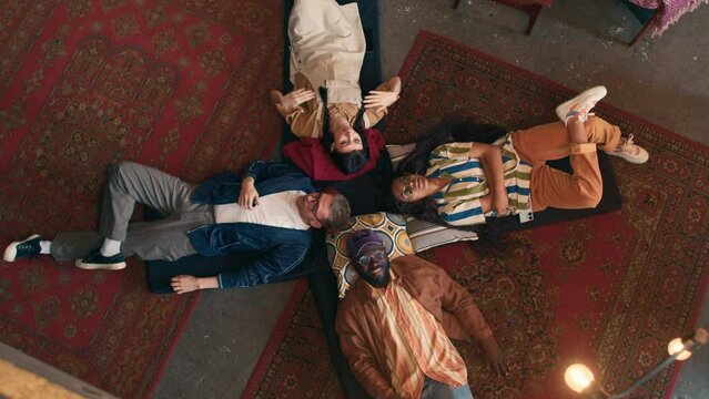 Top-down shot of relaxed young men and women squatting in abandoned house lying on mats on floor talking about everything