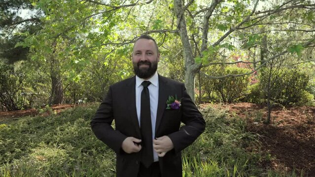 Middle-aged dark-haired unshaven groom putting on black jacket with boutonniere. Smiling groom in wedding suit standing outdoors among trees on sunny day. High quality 4k footage
