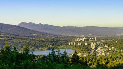 Aerial summer view of Fraser Valley, BC, at Burrard Inlet, featuring high-rise buildings and commercial district of Port Moody and mountain backdrop.
`