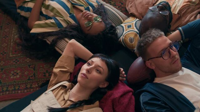 Group of modern young squatters lying on old mats on floor chatting and vaping, 90s aesthetics