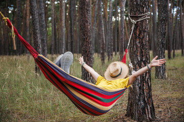 Relaxed woman with covered face by hat and arms outstretched resting in hammock in forest