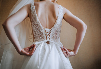 A beautiful bride stands with her back with a knotted white dress corset and a long veil in the room. Wedding photography, portrait.