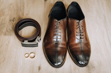Men's brown leather stylish groom's shoes, black belt, gold rings lie on the floor. Close-up wedding photography, top view, set.