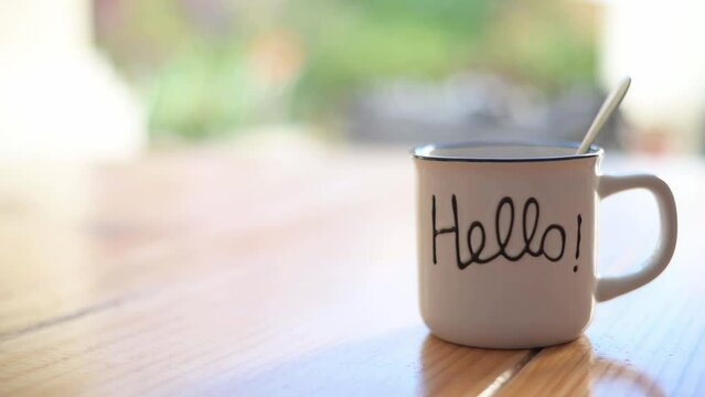 A Hand Pouring Steaming Coffee Into A Cup That Has The Word, Hello, On A Table At Home