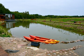 A view of a small pond or lake located next to a wooden path or walkway and a rocky coast with some leisure spot and kayaks visible on a cloudy yet warm summer day on the countryside