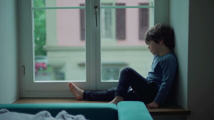 Bored child sitting by window looking at drizzle fall during rainy afternoon. Little boy stuck at home wanting to go out, sad thoughtful expression