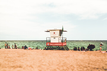 Fototapeta premium Bapselona, Spain. August 1, 2023 White lifeguard tower on sandy beach against green water in a sea. People relaxing on the Mediterranean coast on summer vacation in summertime Tilt shift travel photo.