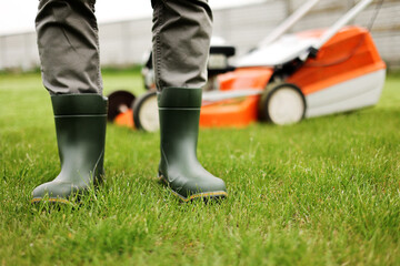 Fototapeta premium Cropped photo of male gardener legs in rubber boots stands on cut green grass lawn at backyard of house. Modern electric cordless lawnmower on back. Landscaping industry theme.