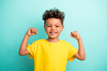 Portrait of good mood strong small boy with curly hair wear stylish t-shirt clenching fists isolated on turquoise color background