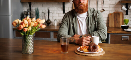 Banner image of bearded and tattooed man holding coffee near fresh pastry and tulips in kitchen 
