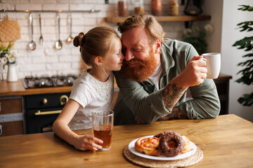 Smiling child kissing father with coffee near sweet pastry during breakfast in kitchen 