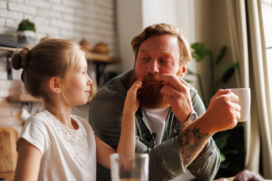 Child touching beard of tattooed father holding coffee and spending time together in kitchen in morning