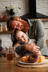 Portrait of cheerful bearded father hugging freckled daughter near blurred pastry and juice in...