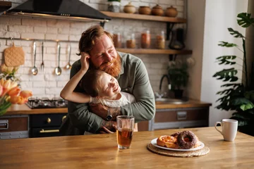 Tuinposter Cheerful daughter hugging bearded father near sweet breakfast in kitchen in morning © Dmytro Hai