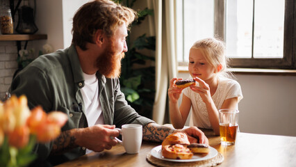 Child holding donut near tattooed father with coffee during breakfast in kitchen in morning