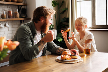 Positive child holding donut and talking to tattooed father with coffee in kitchen 