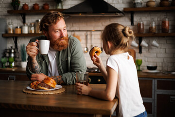 Tattooed and bearded father holding coffee while daughter holding donut during breakfast in kitchen 
