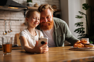 Smiling freckled child taking selfie on smartphone with bearded father near pastry in kitchen 