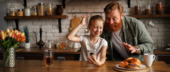 Fototapeta na wymiar Banner image of cheerful bearded father and preadolescent daughter using smartphone together near pastry in kitchen 