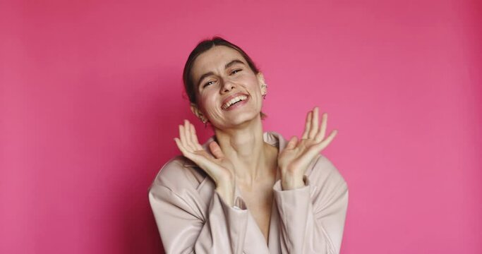 Positive smiling young woman peeking through fingers with curious nosy expression, spying secret information standing on pink background. She open face and laughing. Girl look happy.