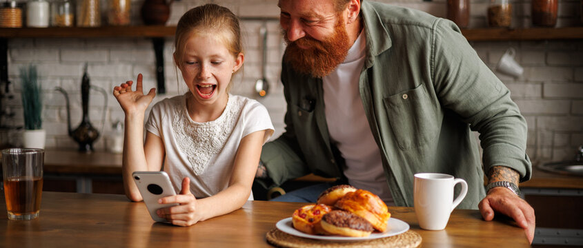 Banner image of preadolescent girl using smartphone with bearded father during breakfast in kitchen