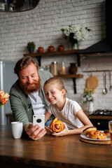Cheerful freckled child holding sweet pastry near father using smartphone in kitchen in morning