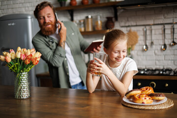 Cheerful freckled girl holding juice near sweet buns near busy parent in kitchen in morning