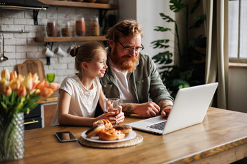Positive girl using laptop together with bearded father during breakfast near smartphone and pastry in kitchen  - obrazy, fototapety, plakaty