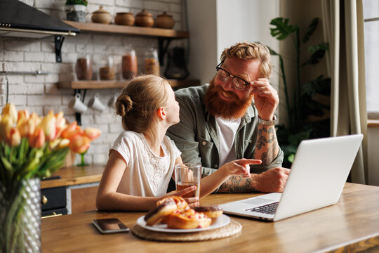 Smiling Tattooed Father Talking To Daughter Pointing At Laptop During Breakfast In Kitchen In Morning