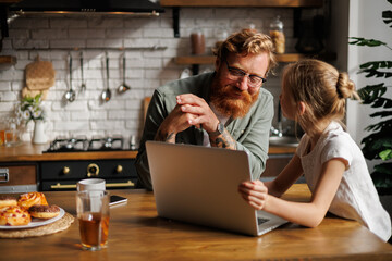 Father and daughter talking near laptop, pastry and drinks in kitchen