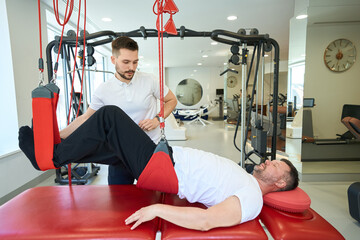 Male doing exercise on fitness equipment assisted by physical therapist