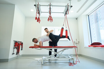Man working out with suspended straps supported by kinesiotherapist
