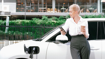 Progressive businesswoman leaning on electric car and charging station before driving around city...
