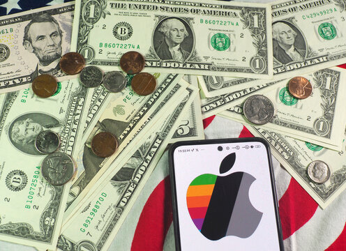 In this photo illustration,  Apple Inc. logo is seen displayed on a smartphone and US currency notes and coins in the background.