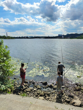 A family of father, mother and son is standing on the bank of a wide river and is fishing with fishing rods. Rear view