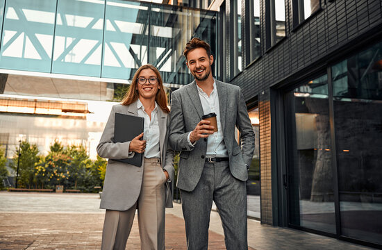 Creative Modern Stylish Confident Elegant Beautiful Successful Cheerful Carefree Business Woman And Man Standing Outside Business Building Holding Laptop And Coffee And Looking At Camera Smiling.