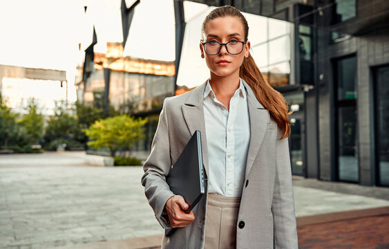 Portrait of beautiful confident serious woman leader stylishly dressed in formal wear and glasses holding laptop and looking at camera. The business center is behind.