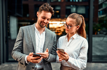 Two adult successful confident business people are standing outside the office holding phones. Attractive bearded man shows something on the phone to a colleague. Project managers, online applications