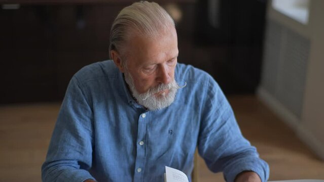 High-angle view of focused mature adult bearded man flipping through pages of book, searching for reference information for job. Caucasian senior male and flips pages of textbook, slow motion.