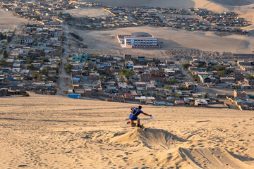 A boy sandboarding as the sun sets over a city in Peru surrounded by sand dunes seen below.