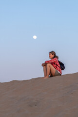 Girl sitting on sand dune with full moon behind her