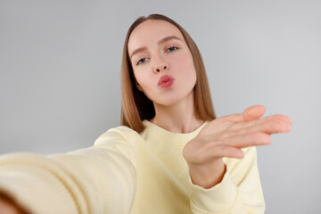 Young woman taking selfie and blowing kiss on light grey background