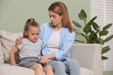 Mother applying ointment onto her daughter's knee on couch indoors