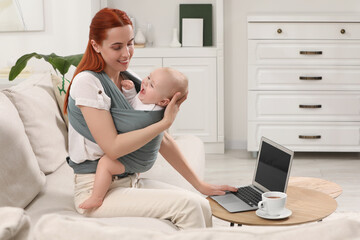 Mother holding her child in sling (baby carrier) while using laptop at home
