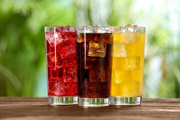 Glasses of different refreshing soda water with ice cubes on wooden table outdoors