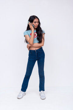 A pensive asian trans woman trying to call someone on the phone, waiting for them to pick up. Wearing a light blue printed shirt and jeans. Whole body photo on a white background.